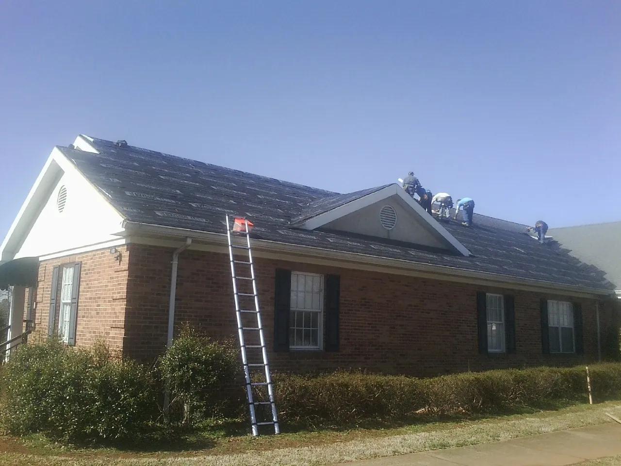 Crew tearing shingles off a brick ranch roof with a ladder set — Emergency Roof Repair work by our crew in Pascagoula