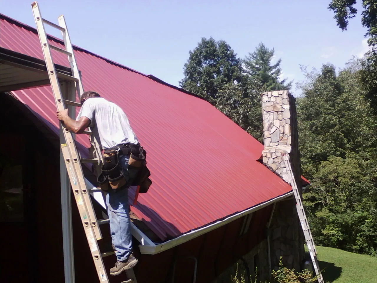 Roofer on a ladder beside a red metal roof and stone chimney — Soffit & Fascia Repair by Roofing Brothers in Pascagoula